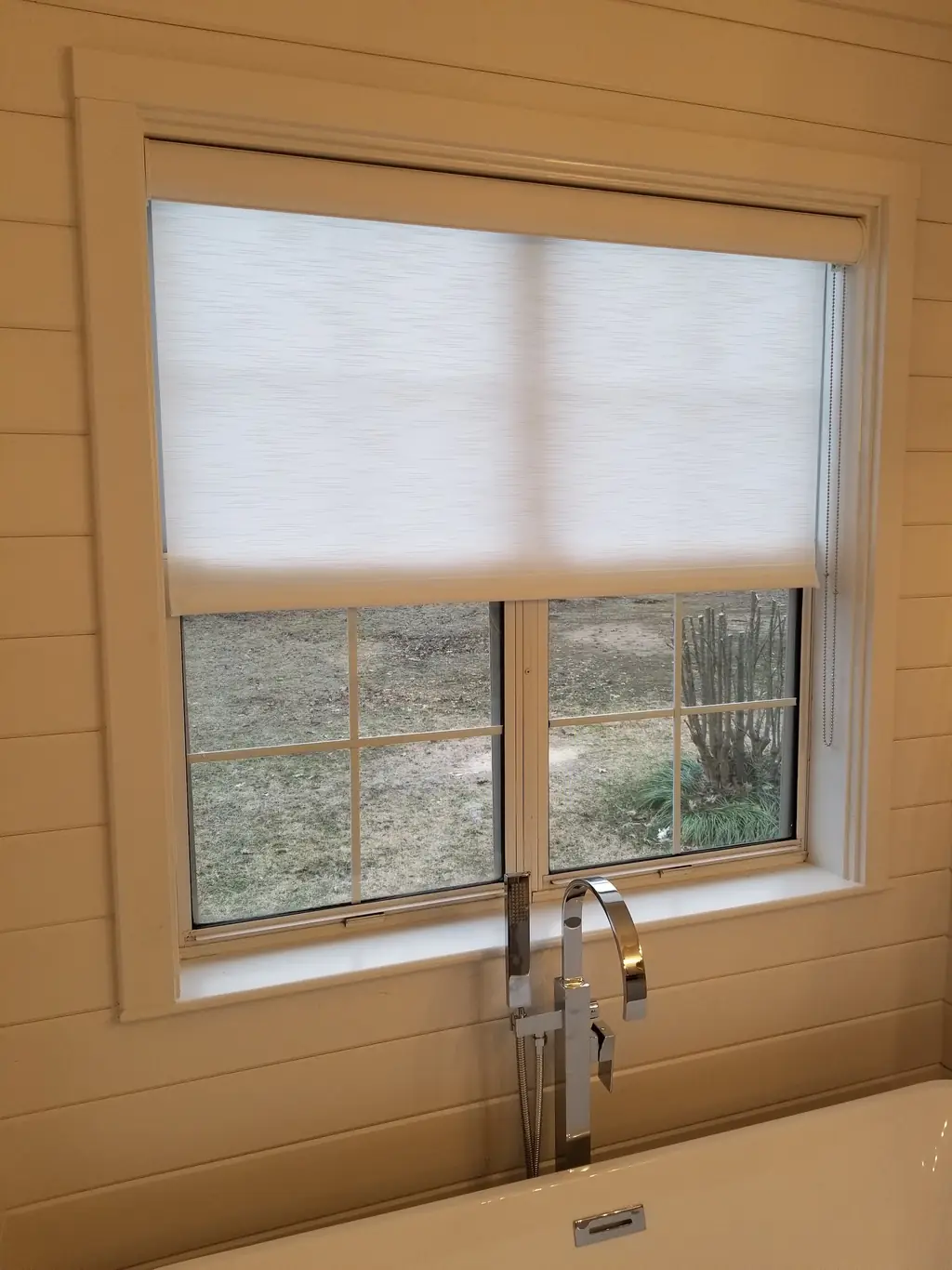 A white roller shade above a freestanding soaker tub in a bright bathroom